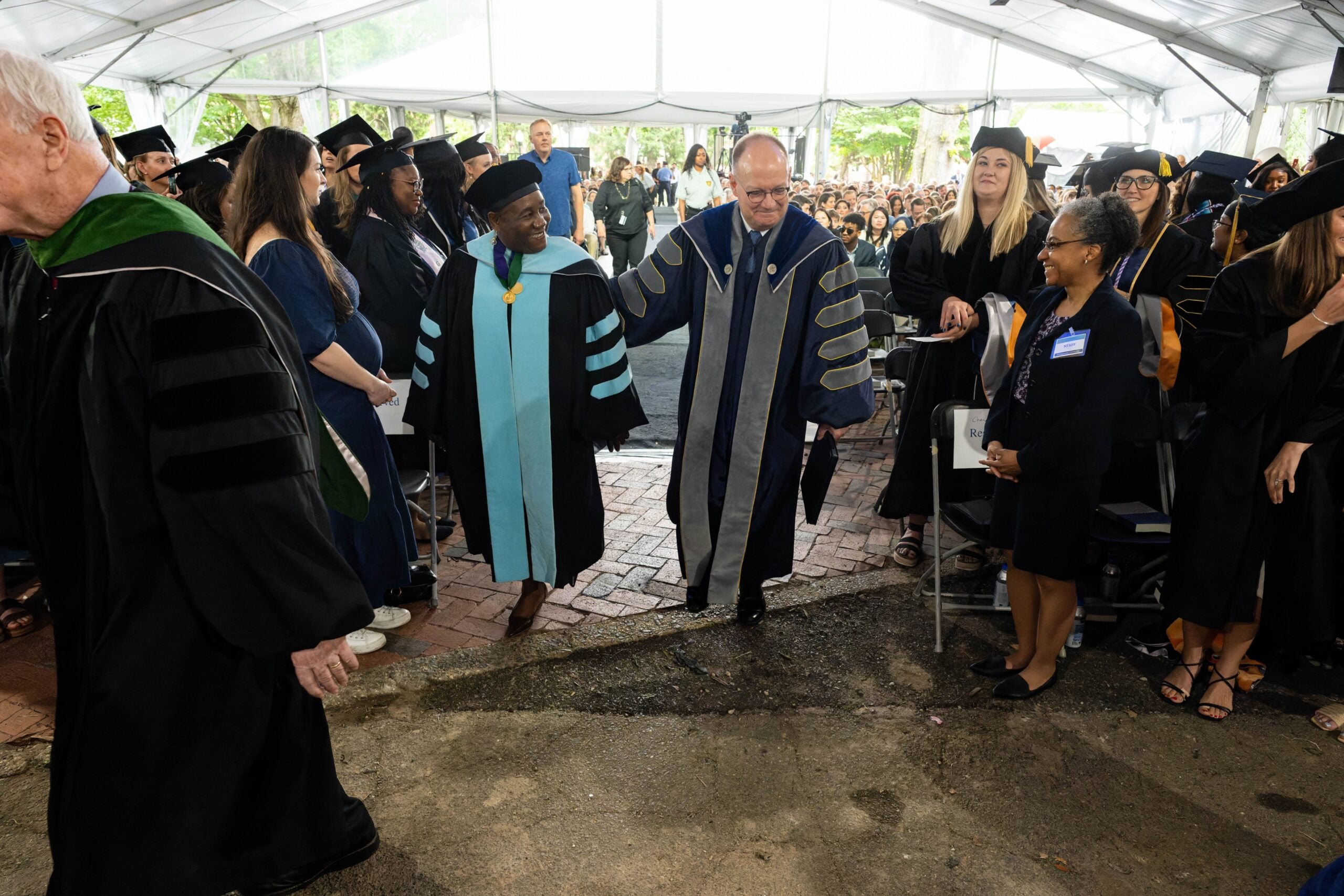 Dr. Roberta Waite and President DeGioia, both wearing academic regalia, walk through a crowd of graduates and guests in an outdoor tent during commencement.