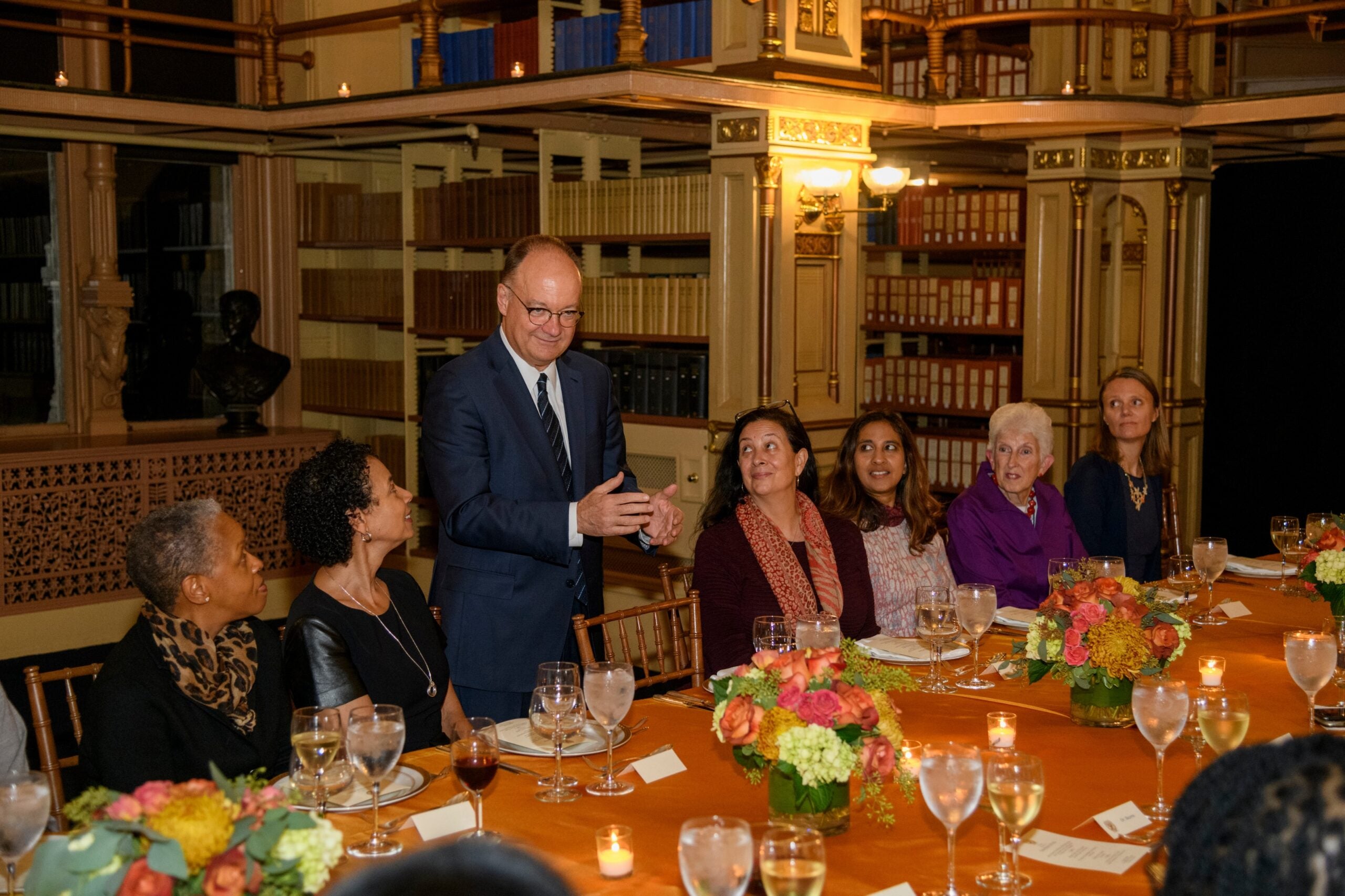 President DeGioia stands with a group of women seated at a formal dinner. They are surrounded by tall bookcases.
