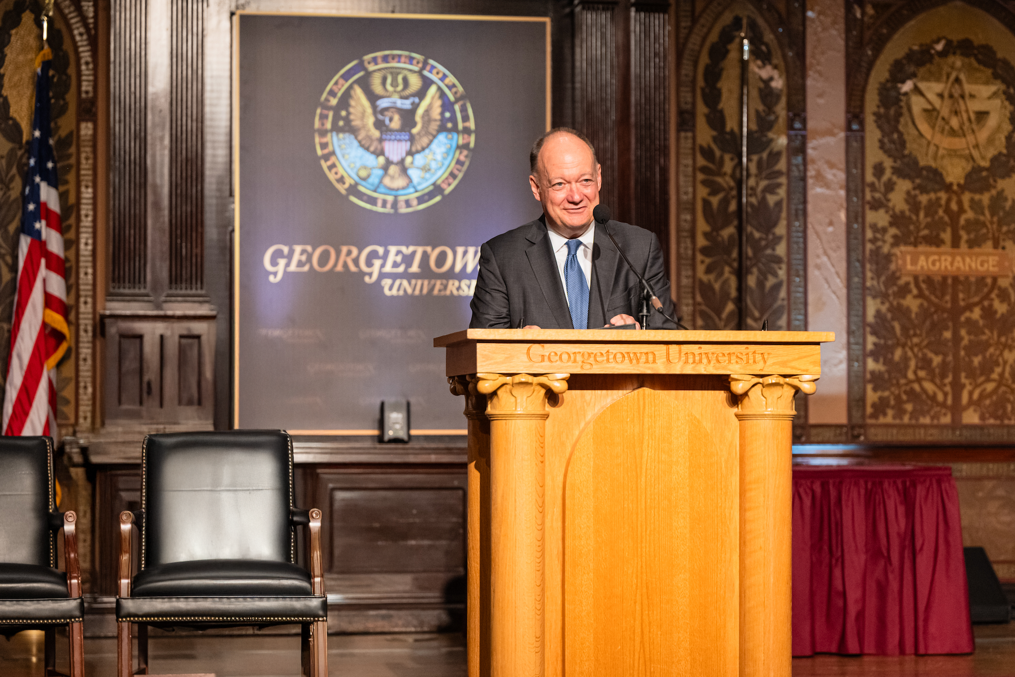 President DeGioia, wearing a suit and tie, speaks from behind a wooden podium. He is in an ornately decorated room with the Georgetown name and seal behind him.