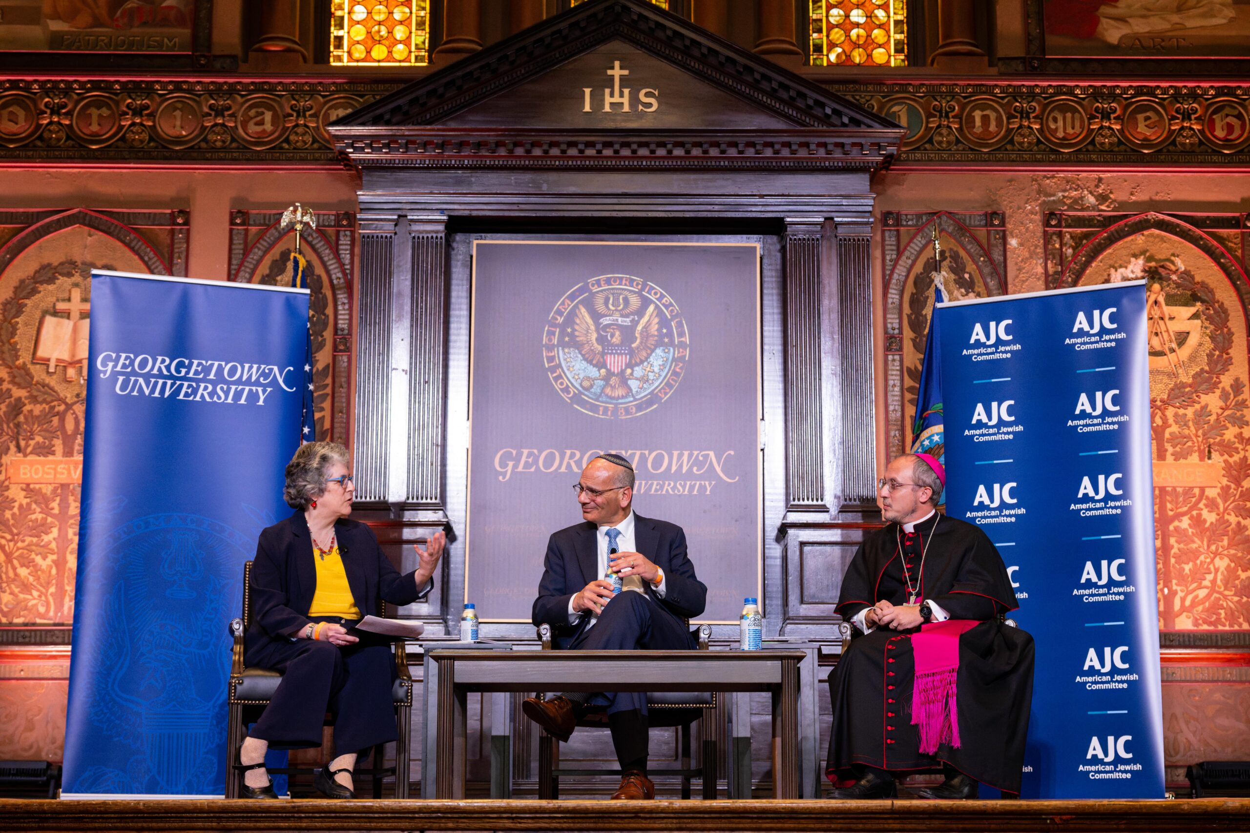 A woman, a Jewish man wearing a yamaka, and a Catholic man wearing Catholic vestments, sit in conversation on a Georgetown University stage.