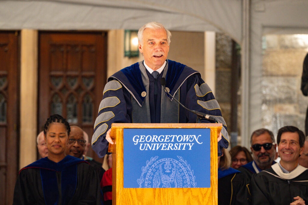 Interim President Robert Groves, wearing academic regalia, speaks from a Georgetown University branded podium. Faculty members are seated behind him.