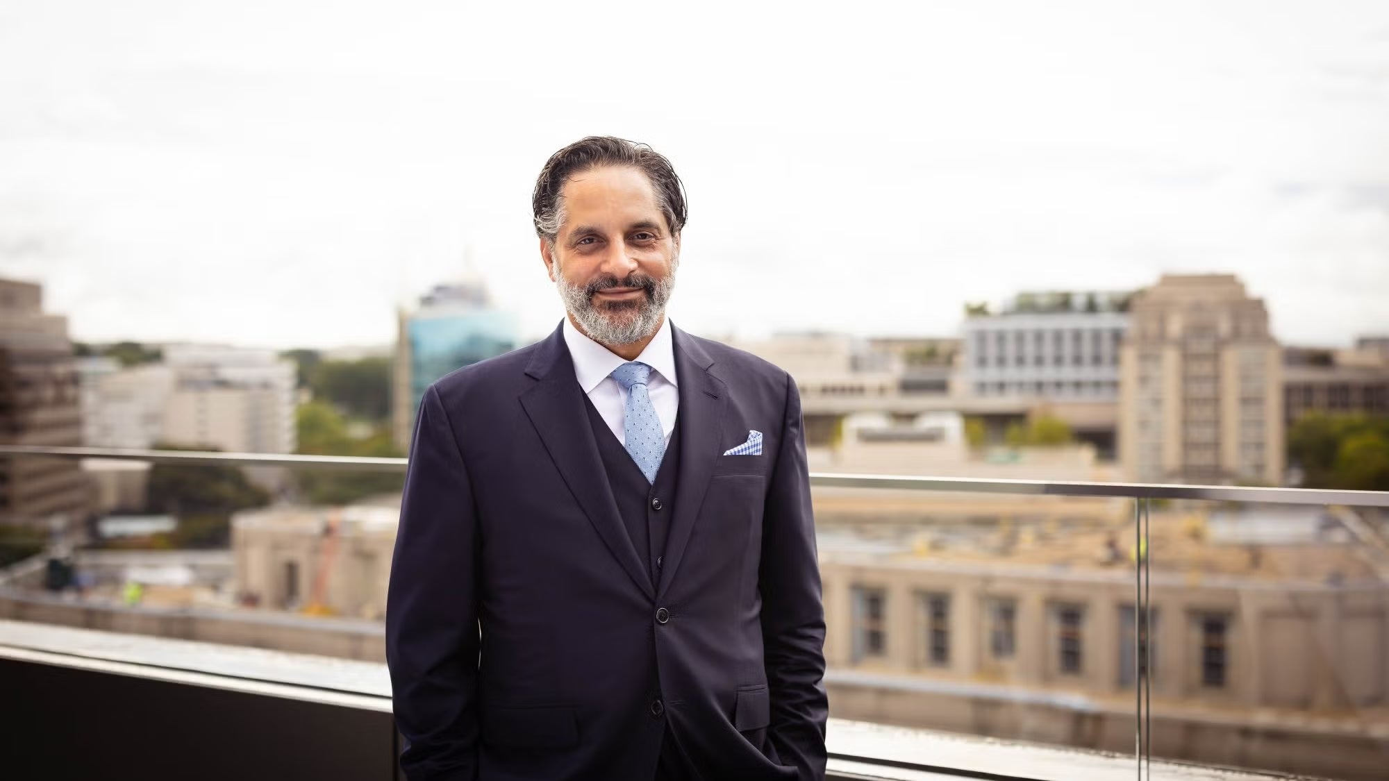A Latino man with dark hair and facial hair, wearing a suit and tie, stands on a balcony overlooking city buildings.