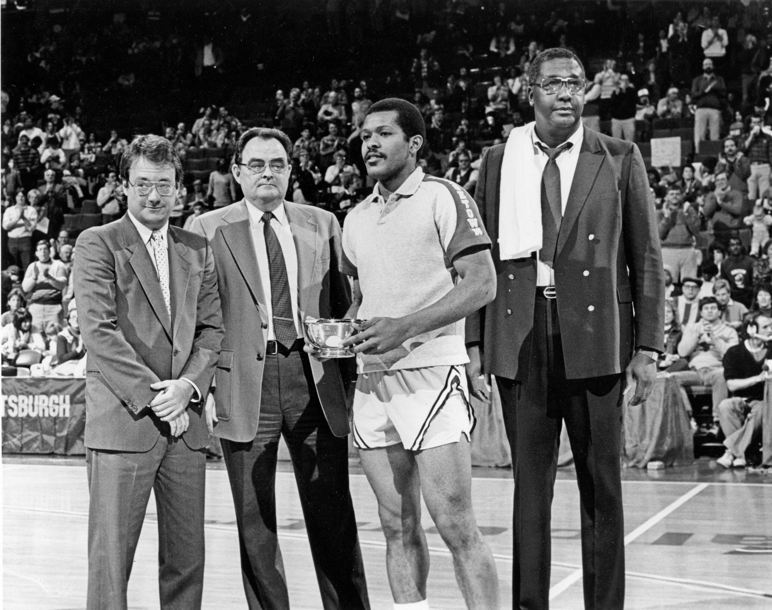 A black and white photo of President John J. DeGioia standing on a basketball court with a player holding a trophy and the late Coach John Thompson Jr.