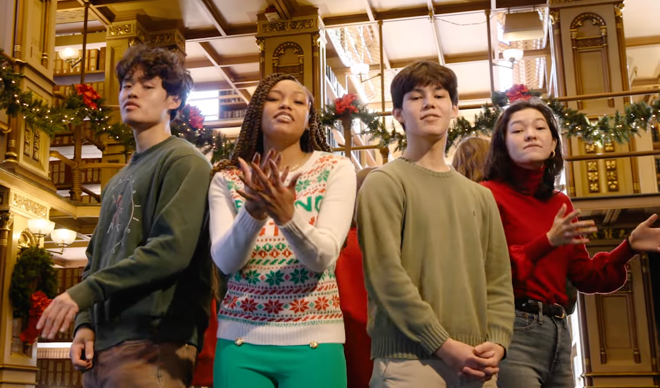 A group of four students in singing in the middle of a library decorated for Christmas.