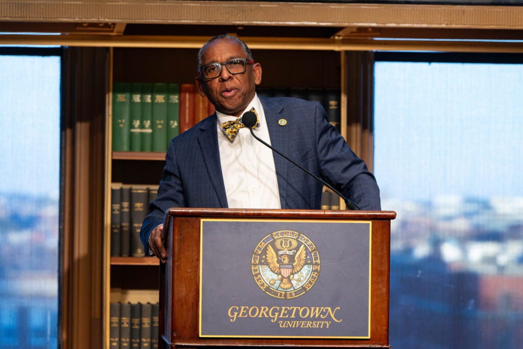 Reynold Verret, a black man wearing a suit and bow tie, speaks from behind a podium in Riggs Library.