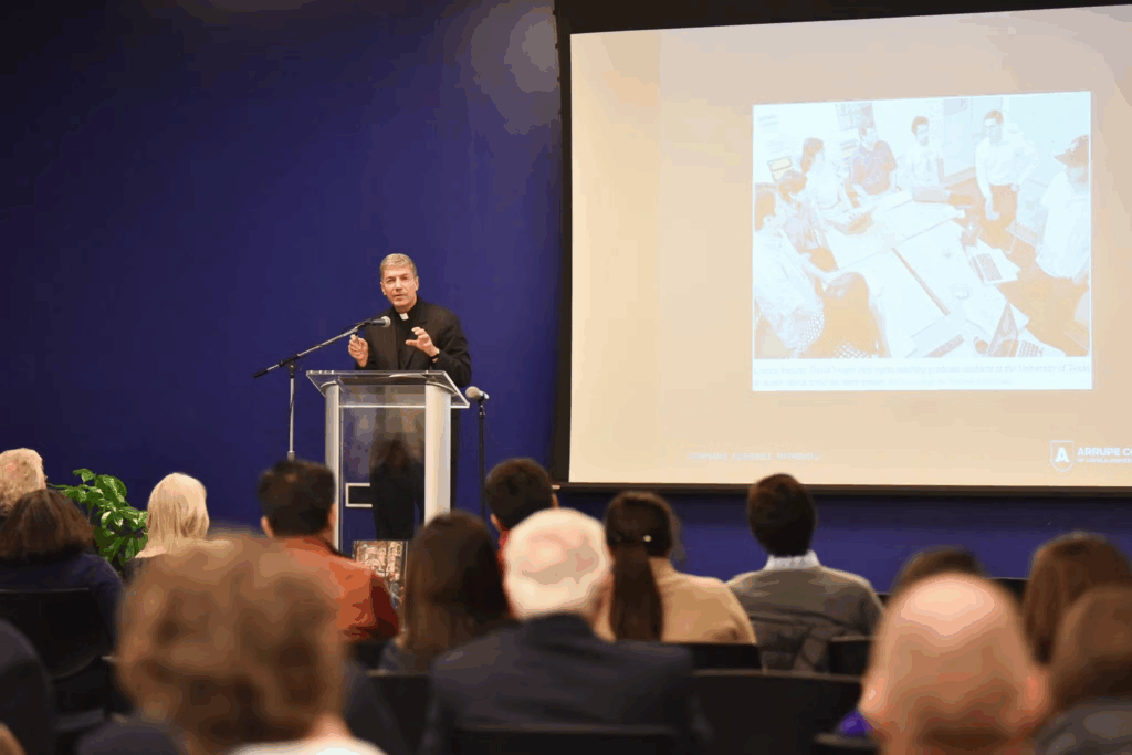 Rev. Steve Katsouros, S.J., a white man in Catholic vestments addresses a seated audience from behind a podium.