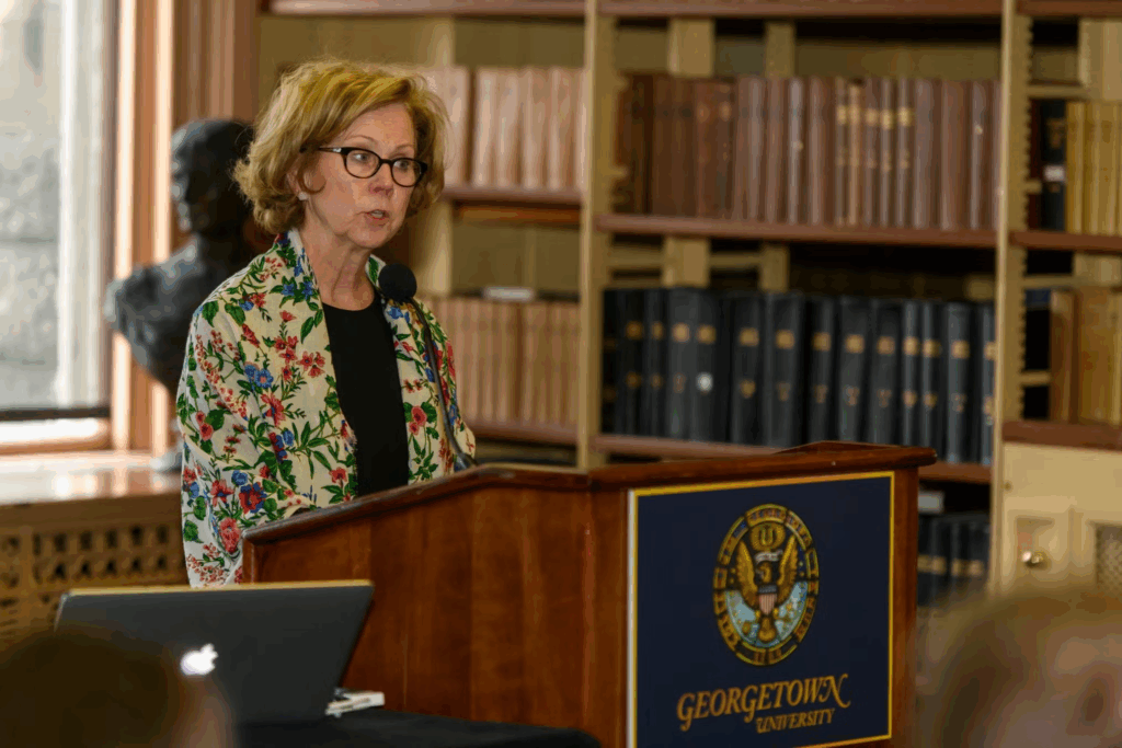 Jeanne Lord, a white woman with short blonde hair and glasses, speaks from behind a podium in Riggs Library.