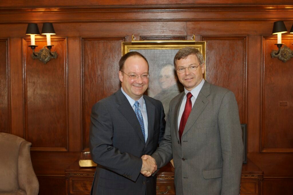 Presiden tJohn J. DeGioia and Prime Minister Kjell Magne Bondevik, both older white men wearing suits, pose while shaking hands. Behind them is a portrait painting.