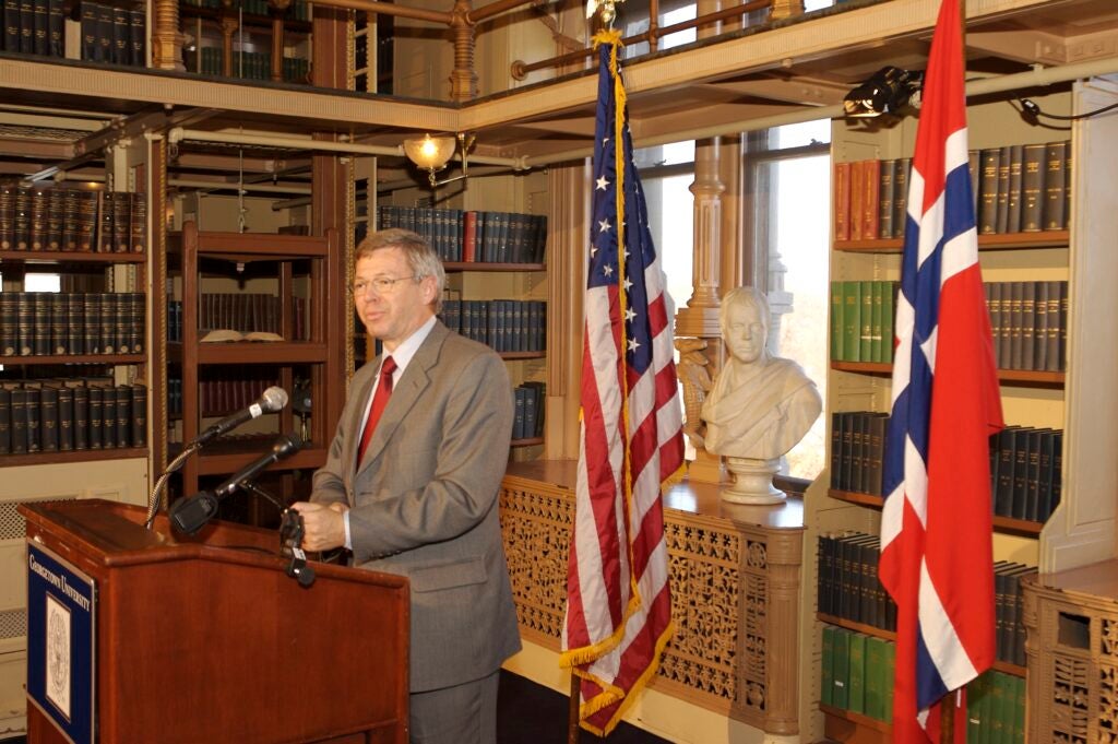Prime Minister Kjell Magne Bondevik, an older white man wearing a suit, speaks from behind a podium in a large library. Behind him are the US and Norwegian flags.