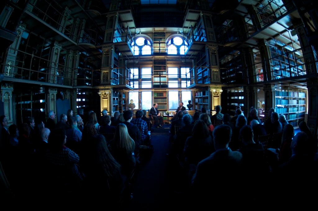 Wide shot of the Riggs Library, showing two men sitting in dialogue in front of a large seated audience.