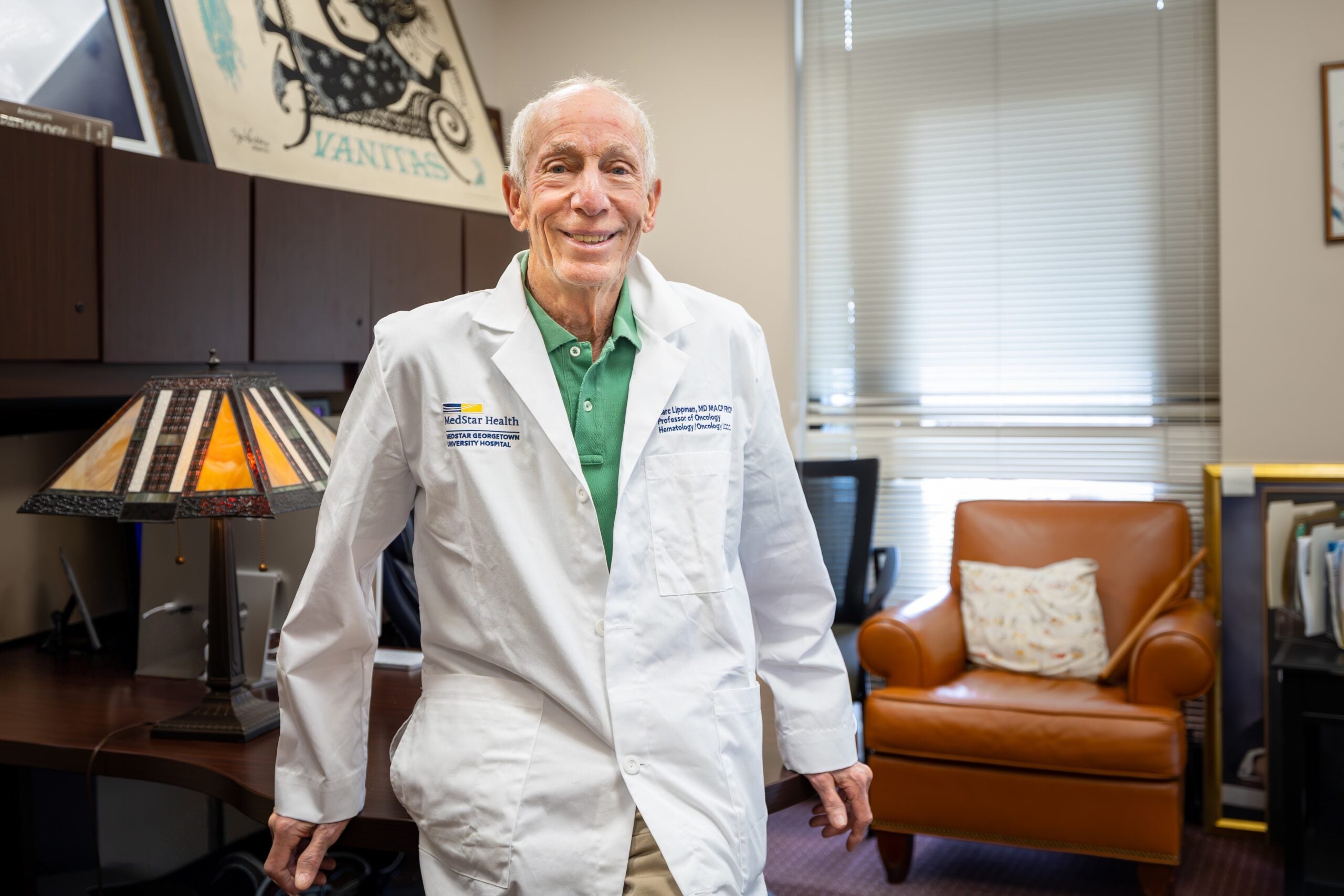 Marc Lippman, an older white man wearing a MedStar Health branded doctor's coat, smiles and poses in front of the desk in his office.