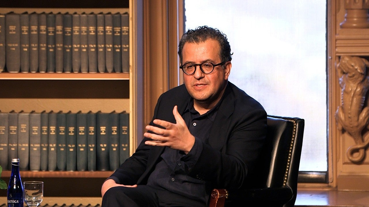 Hisham Matar, an American-Libyan man with dark curly hair and glasses, is seated in Riggs Library, speaking to an audience off-camera.