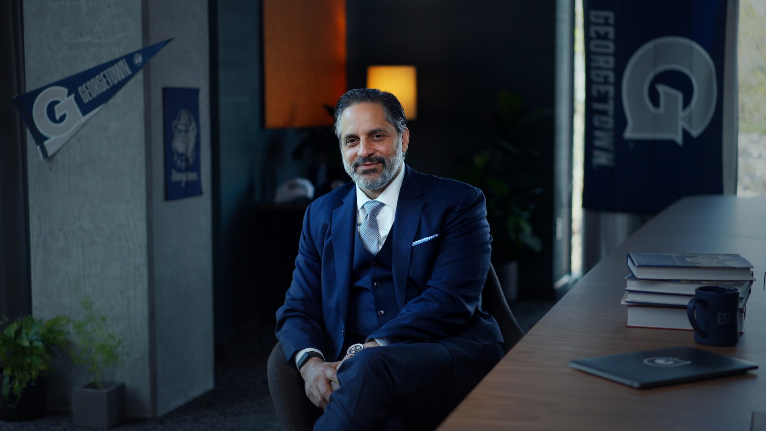 Eduardo Peñalver, a Cuban man with dark hair and facial hair, wearing a navy 3-piece suit, sits at a desk, surrounded by Georgetown University decor.