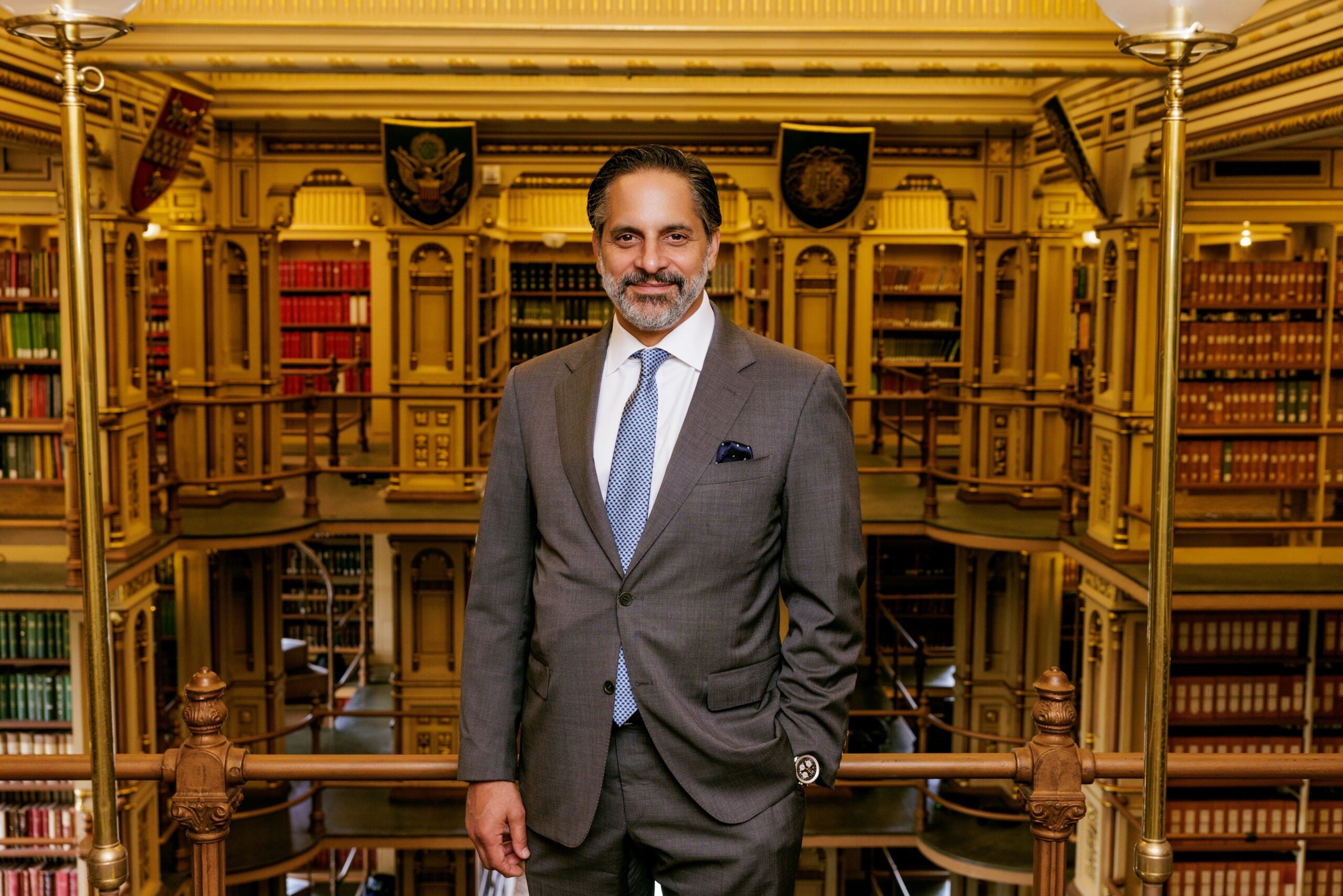 Eduardo Peñalver, a Cuban man with dark hair and facial hair, wearing a medium gray 3-piece suit with a blue tie, stands in front of the library bookshelves in Riggs Library.