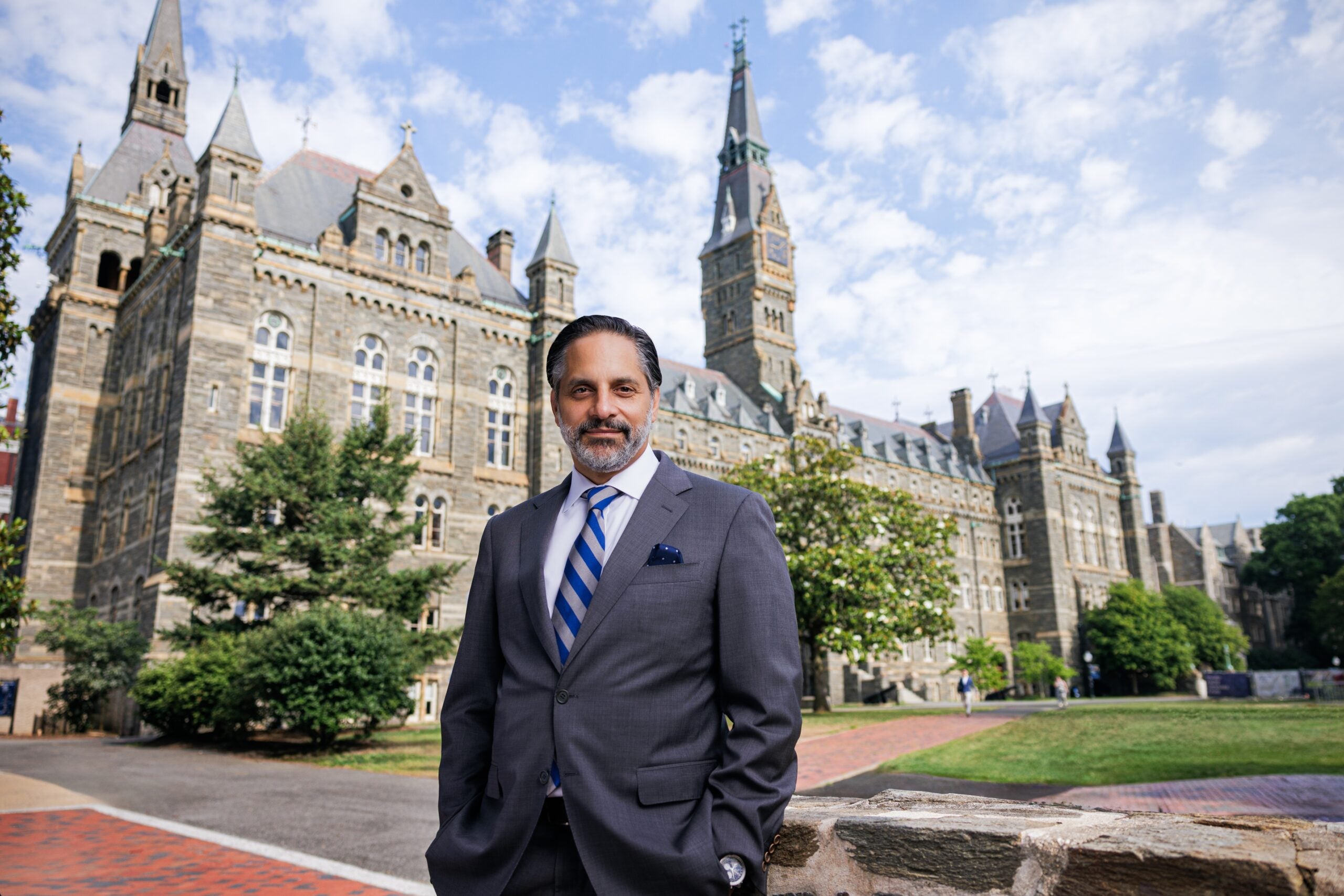 Eduardo Peñalver, a Cuban man with dark hair and facial hair, wearing a gray suit with a blue and gray tie, stands outside in front of Healy Hall.