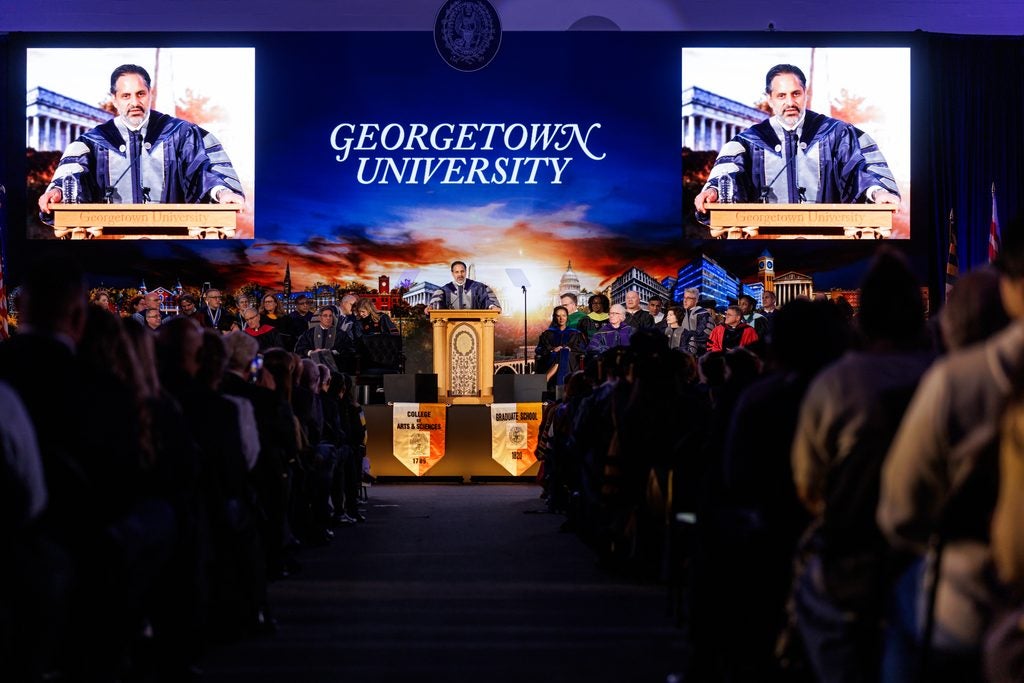 President Peñalver stands behind an ornate wooden podium in front of a large seated crowd. Behind him is a screen projection of the campus at sunset with two inset video close-ups of him.